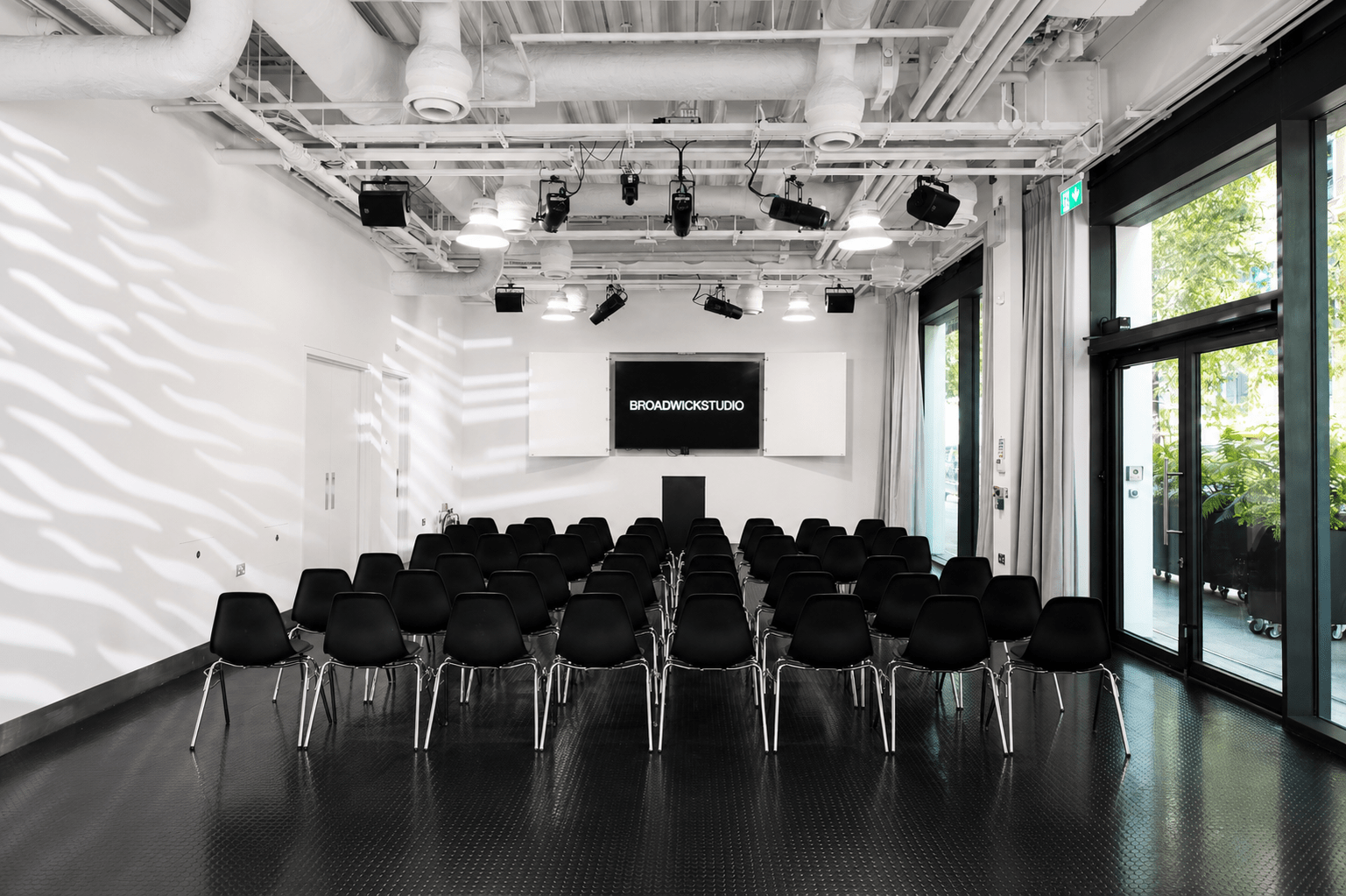 Lecture room with rows of black chairs facing a stage and a screen reading BROADWICKSTUDIO, white walls, and exposed ceiling pipes. Large windows/doors on the right.